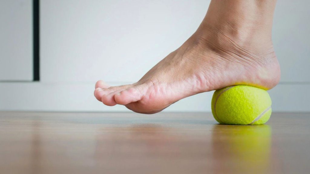 A person's foot applying pressure on a tennis ball, potentially to alleviate foot pain